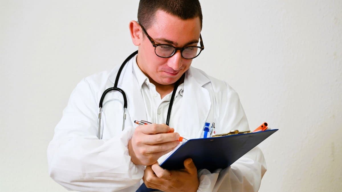 A male doctor in a white coat writes on a clipboard in a hospital setting, showcasing healthcare professionalism.