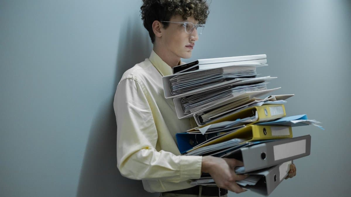 An overworked office worker carrying a large stack of files and folders in a modern setting.
