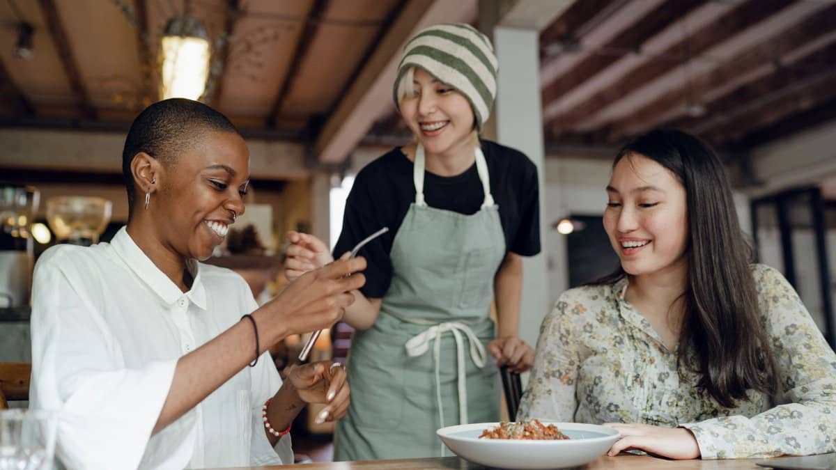 Happy laughing diverse friends in smart casual clothes sitting at table in cozy cafe and eating delicious food near smiling Asian waitress