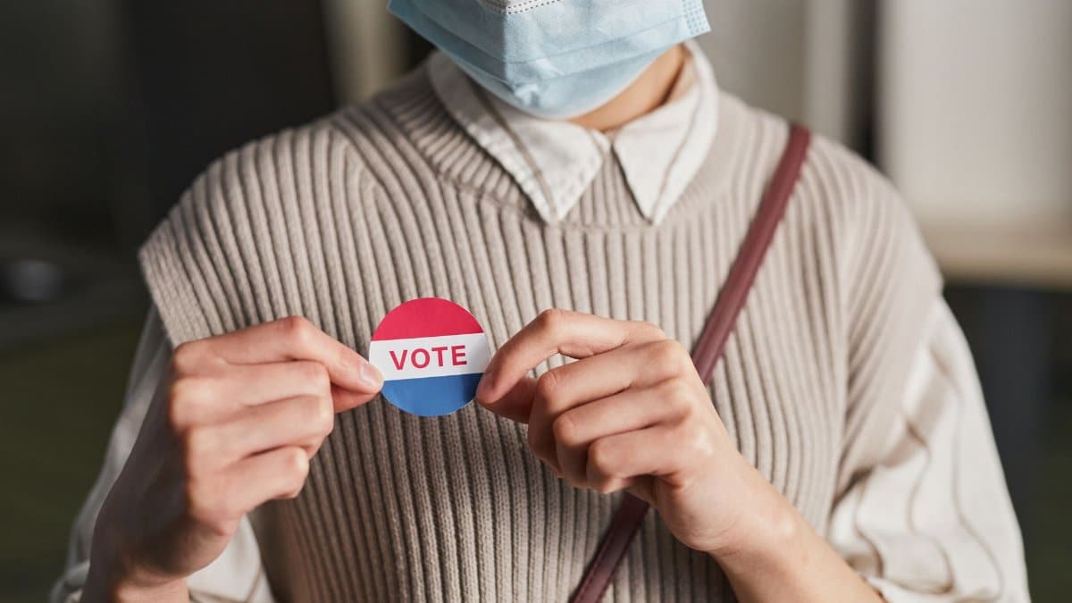 A person wearing a mask holds a voting badge, symbolizing participation in the election.