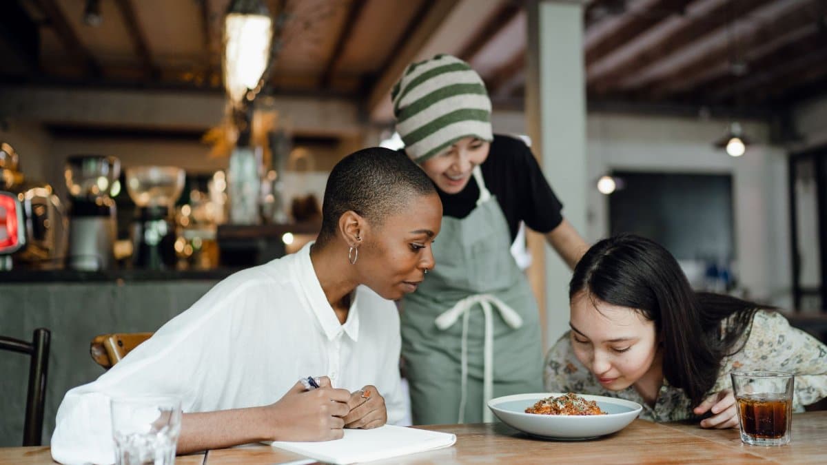 Content multiethnic female restaurant workers sitting at table with notepad while tasting and smelling dish from new menu