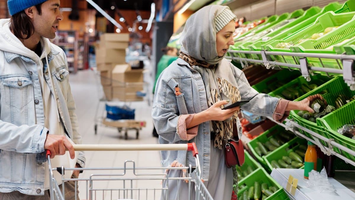 Diverse couple shopping for vegetables in a supermarket, discussing choices with smartphone.