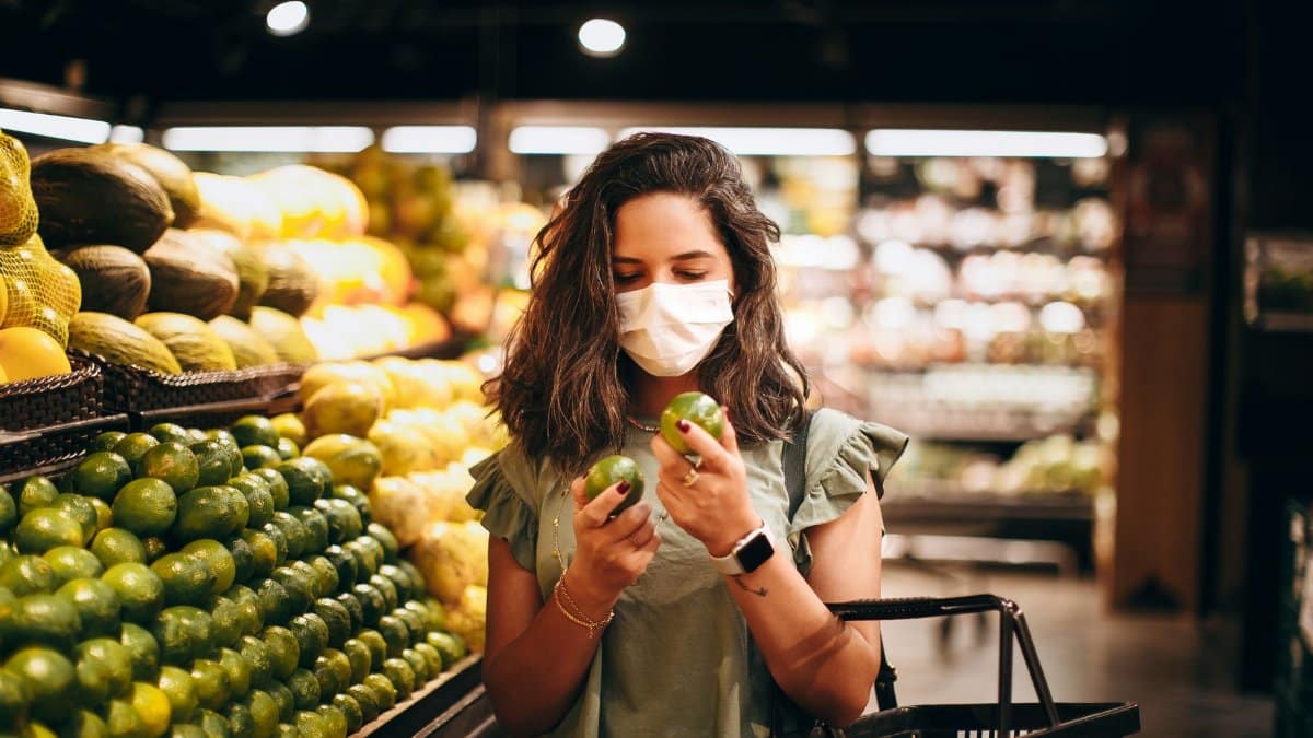 A woman wearing a mask shops for green apples in a supermarket produce section.