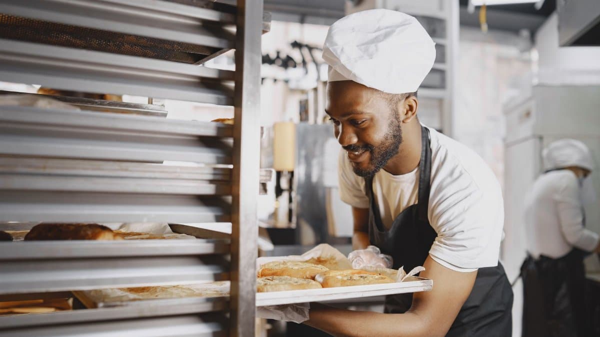 A cheerful baker in a professional kitchen holding a tray of freshly baked breads.