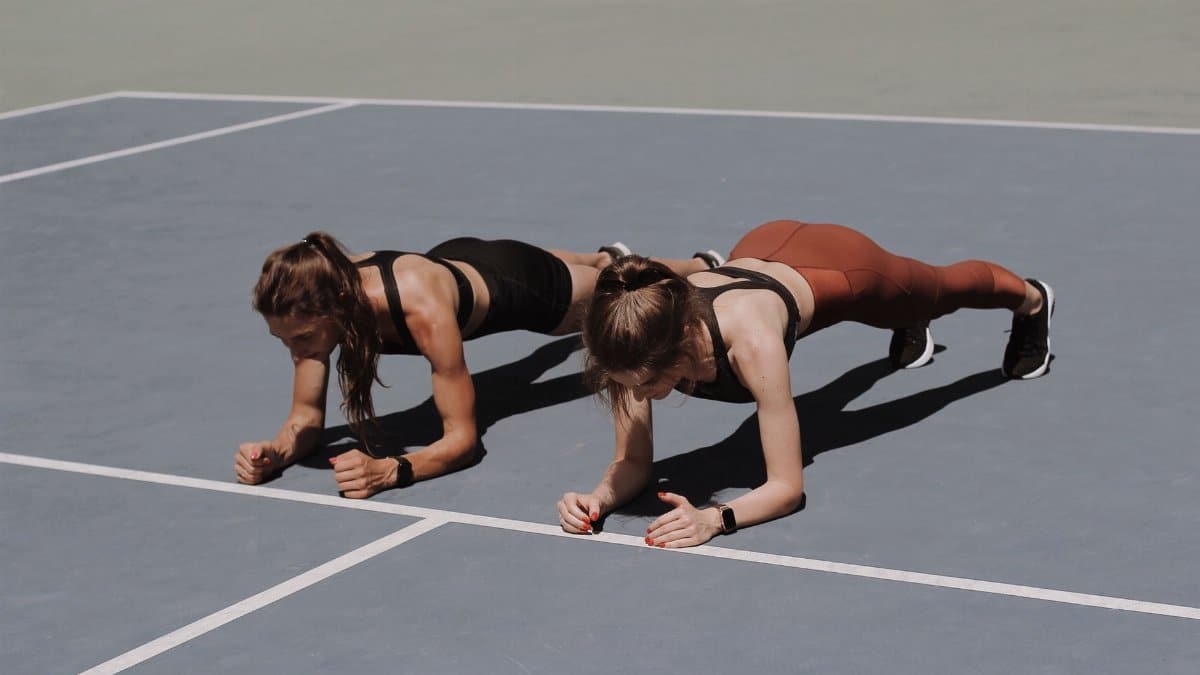 Two women in activewear doing plank exercises on an outdoor sports court.