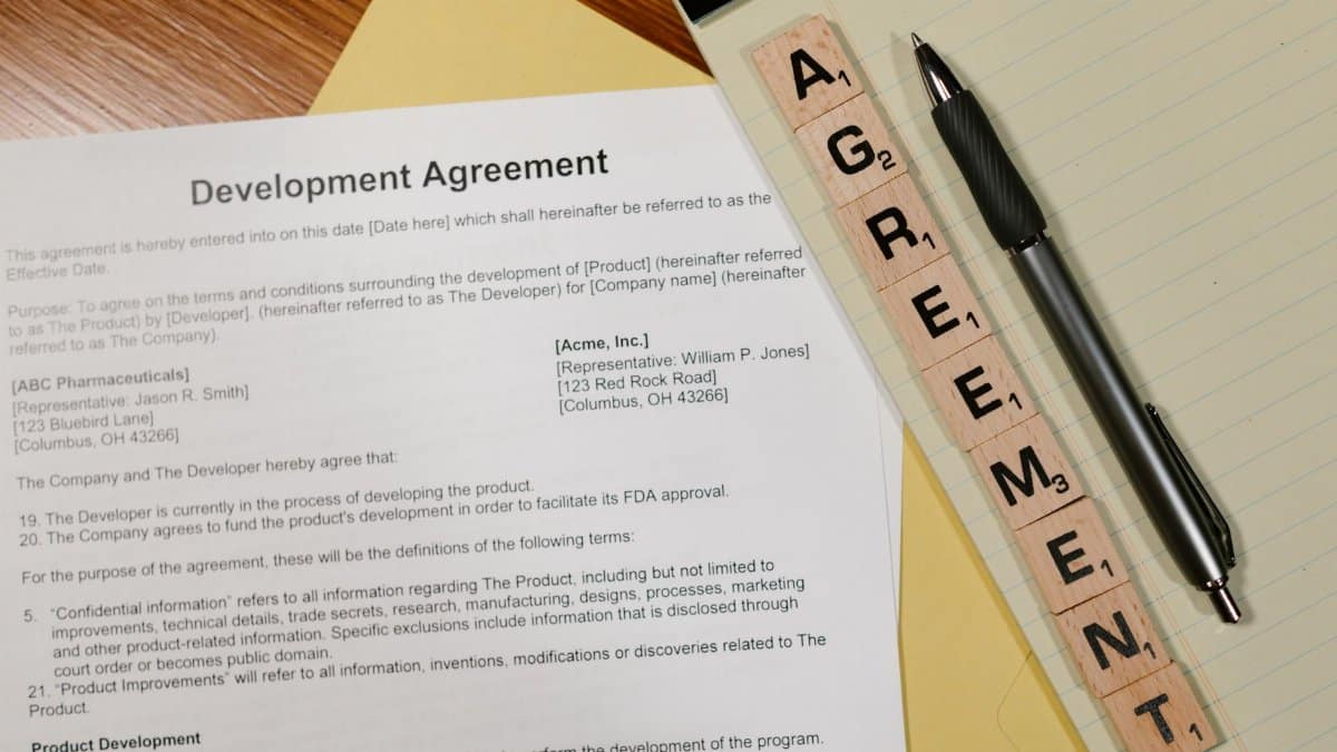 Close-up of a development agreement document with pen and Scrabble tiles spelling 'AGREEMENT.'