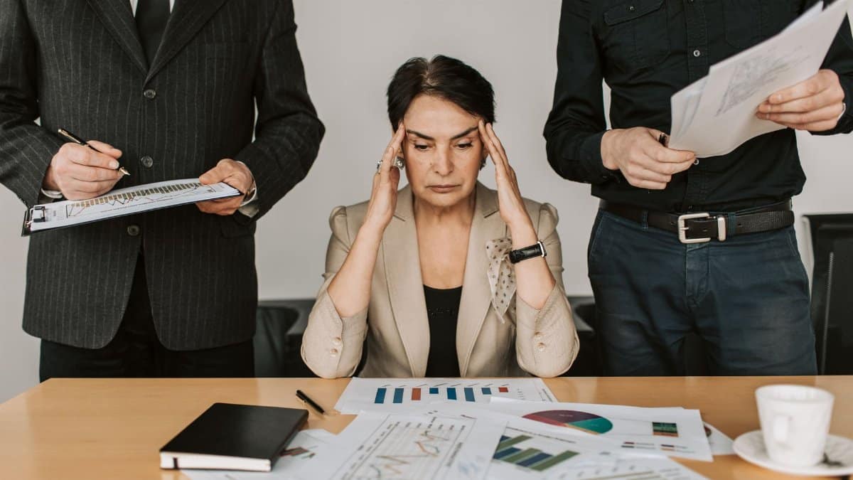 A stressed businesswoman holding her head with colleagues holding documents in a corporate office setting.