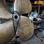 Two men working together to handle a massive industrial propeller in a workshop setting.