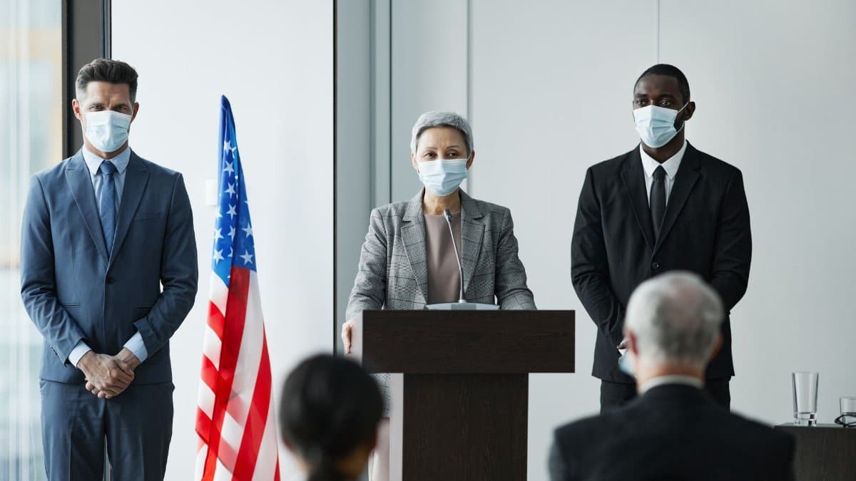 Masked business leaders at a conference podium with US flag backdrop, emphasizing safety and professionalism.