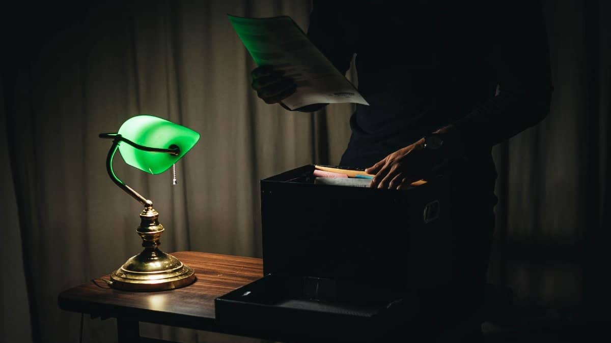 Unrecognizable male with sheets of paper standing near wooden table with glowing table lamp and box of documents in dark room while working late at home
