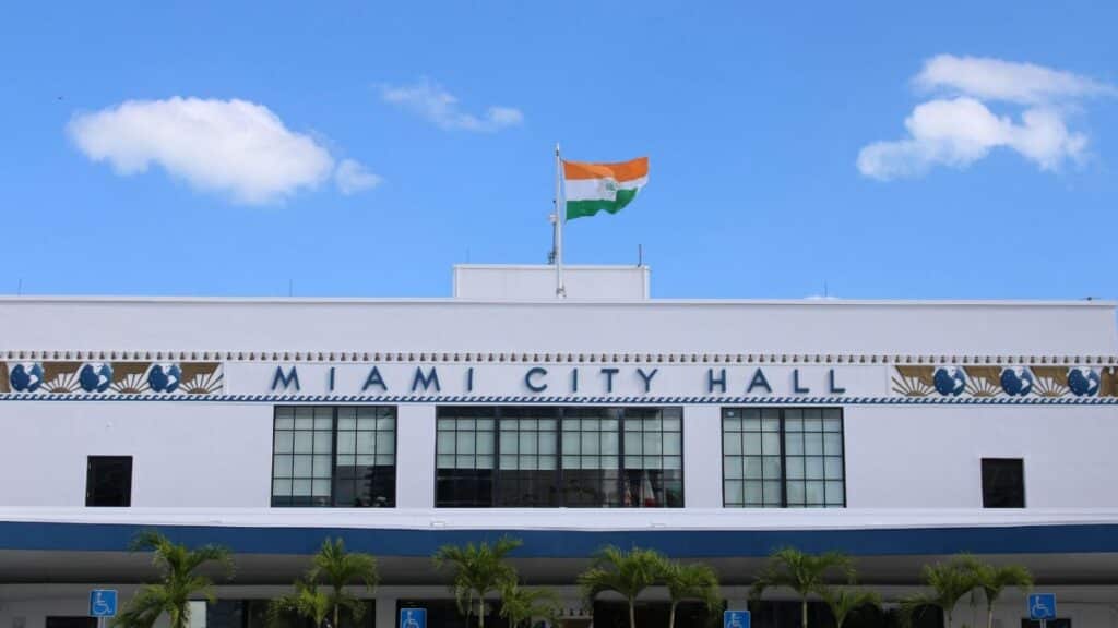 A clear view of Miami City Hall displaying an Indian flag under a bright blue sky.