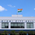 A clear view of Miami City Hall displaying an Indian flag under a bright blue sky.