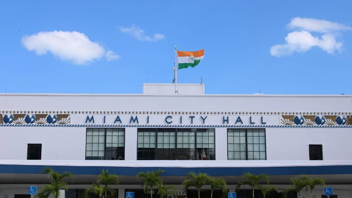 A clear view of Miami City Hall displaying an Indian flag under a bright blue sky.