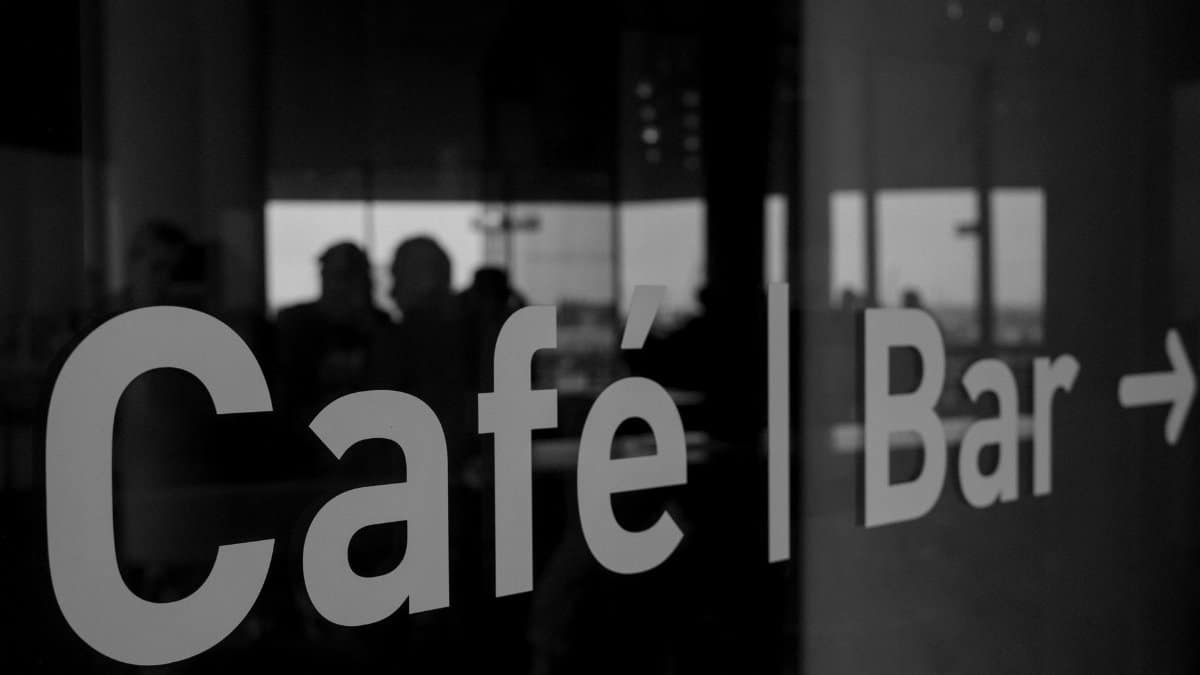 Elegant black and white photo of a café bar sign with people reflected inside, creating a moody atmosphere.