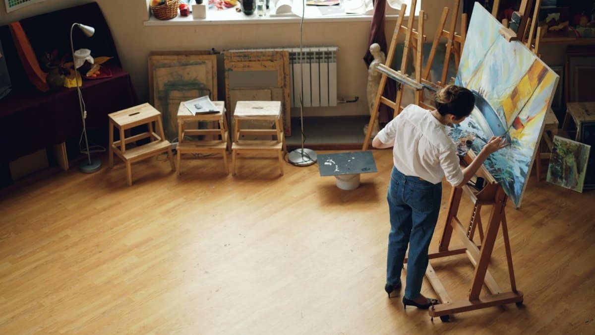 A woman painting on a canvas in a well-lit artist studio filled with creative supplies.