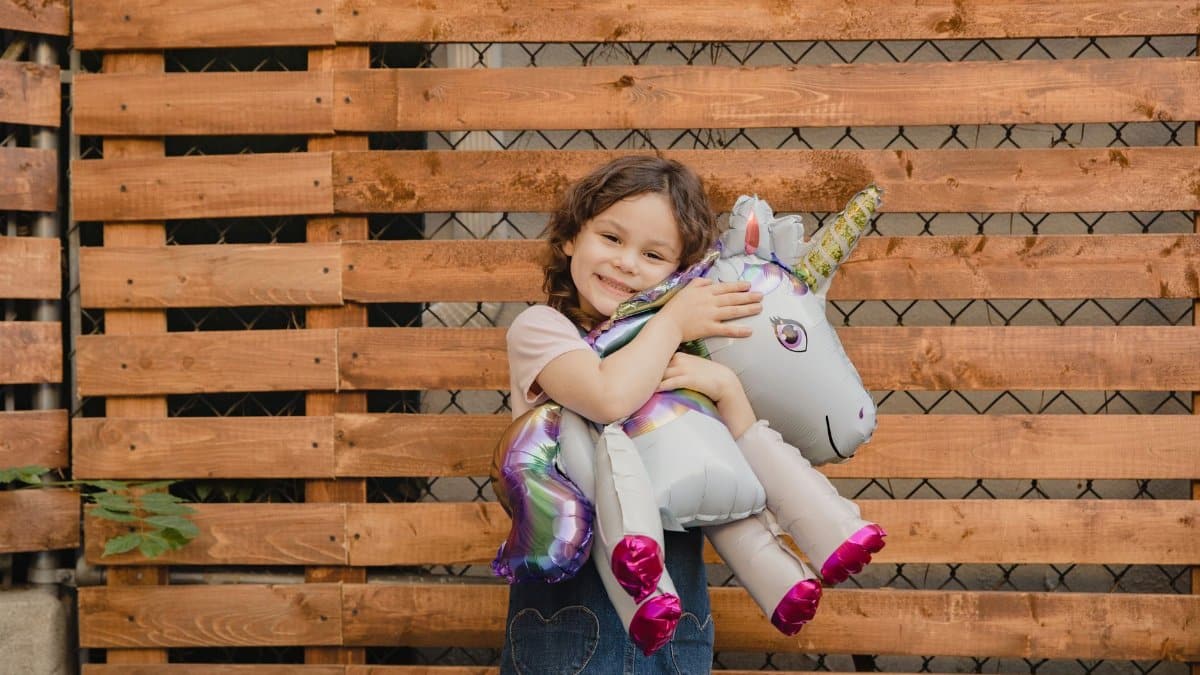 Smiling girl hugging a unicorn balloon against a wooden fence on a sunny day.