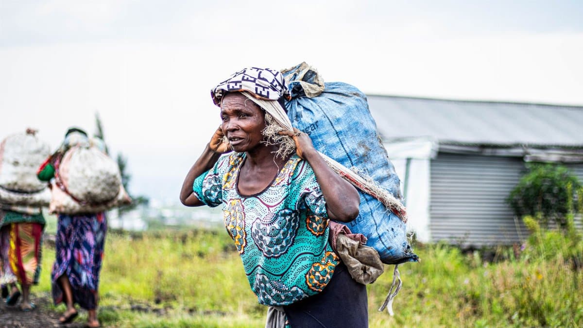 African women walking outdoors carrying heavy loads. Vibrant attire and rural setting.