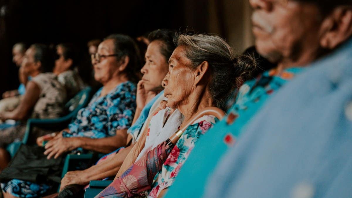 A group of senior adults attentively listening in an indoor auditorium setting.