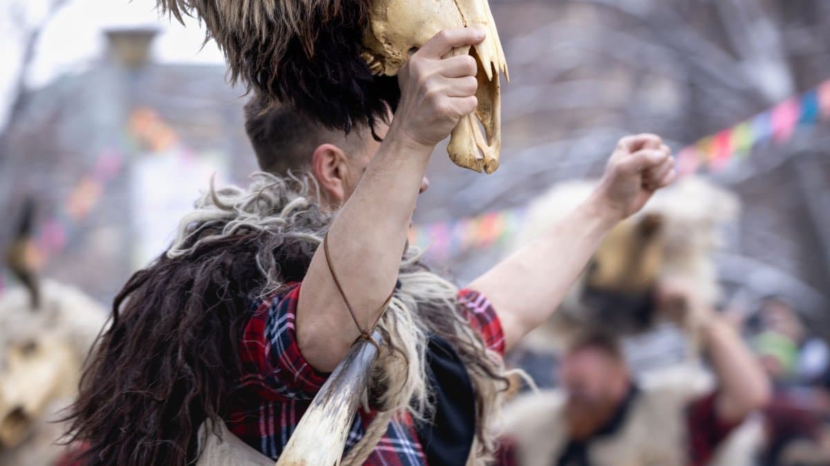 Man in furs and plaid shirt holding animal skull at an outdoor festival, embodying cultural traditions.