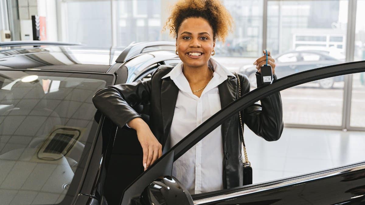 Smiling woman with car keys standing beside her new vehicle in a dealership showroom.