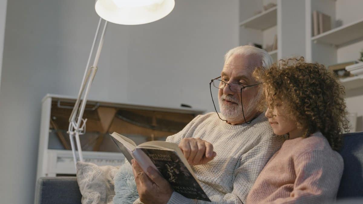 An elderly man reading a book with his granddaughter in a cozy indoor setting.