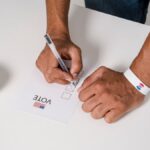 A person wearing a wristband fills out a voting ballot with a pen. Voting stickers visible.