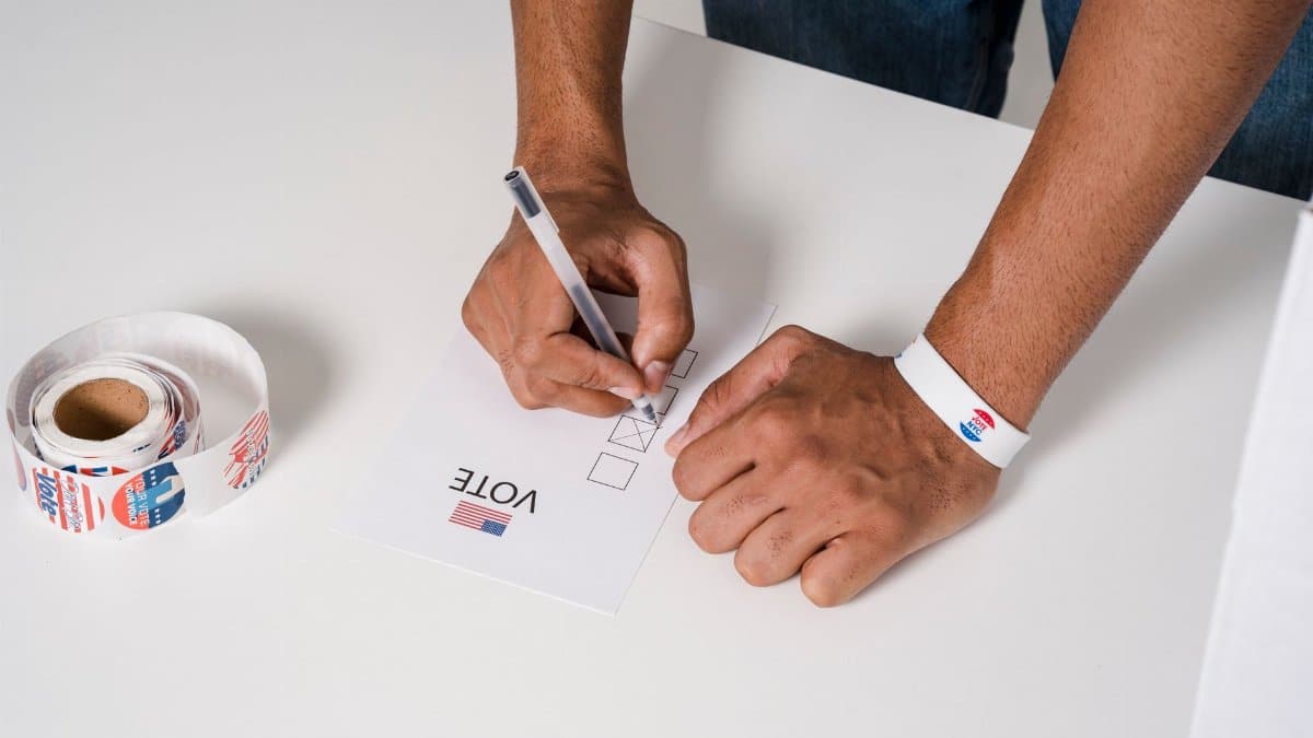 A person wearing a wristband fills out a voting ballot with a pen. Voting stickers visible.