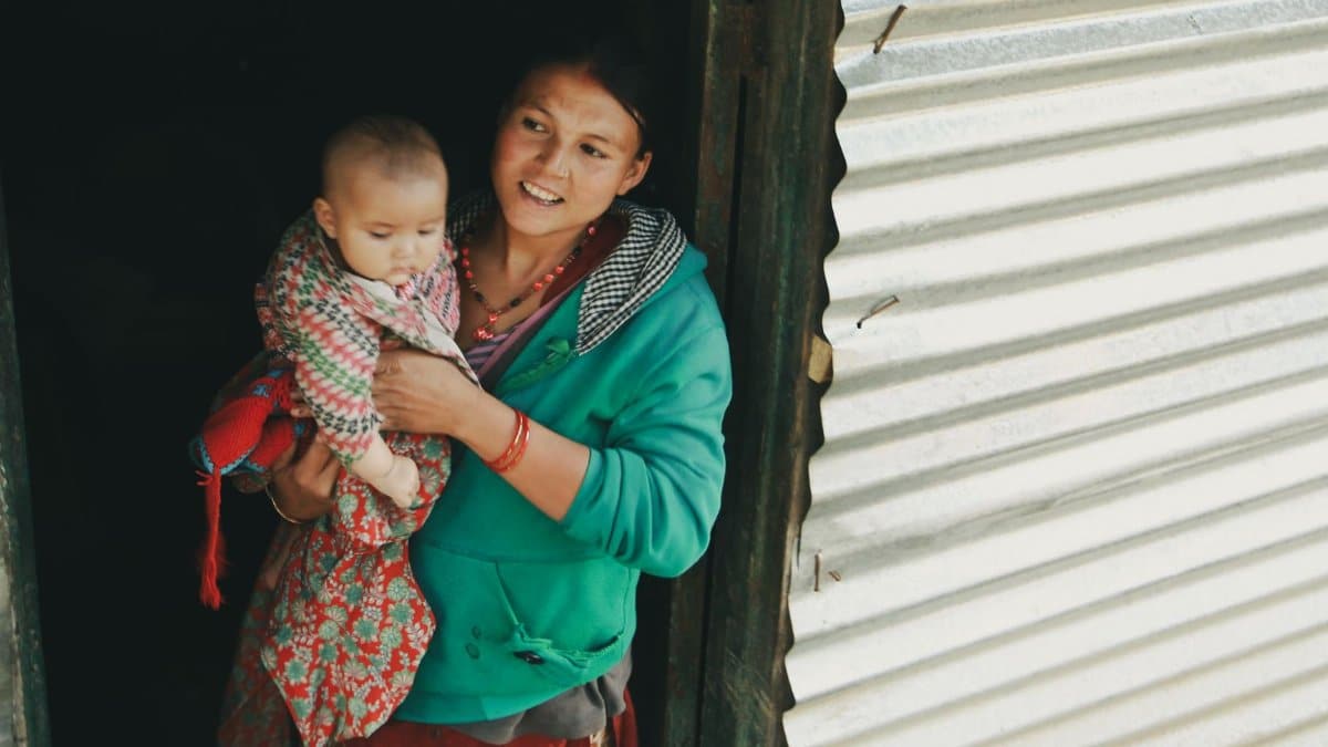 Smiling mother holds her baby while standing at the doorway of a rustic home, showcasing familial bond.