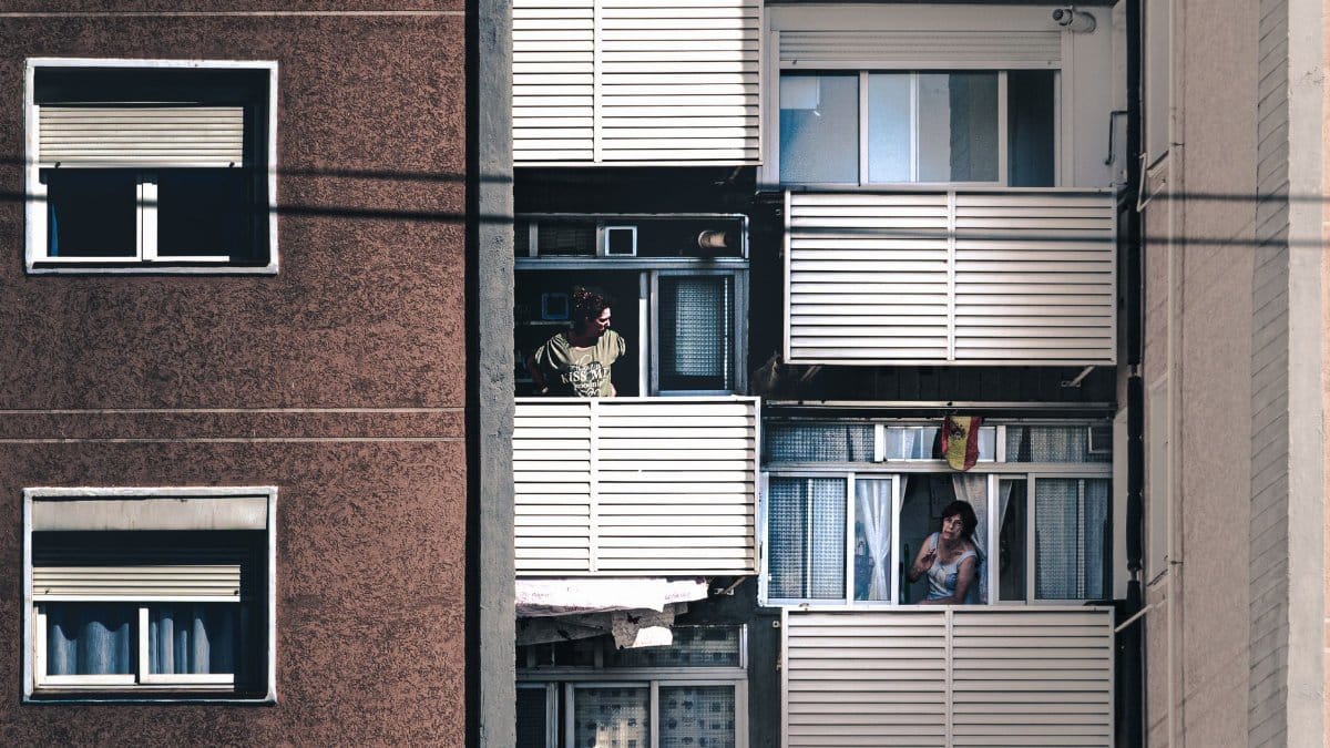 Two people standing on apartment balconies in a city building, interacting.