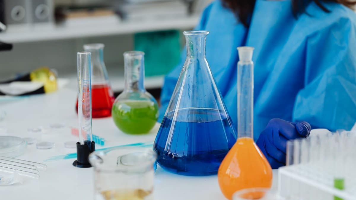 A close-up of a laboratory desk with colorful chemicals in glassware, featuring a researcher in protective gear.
