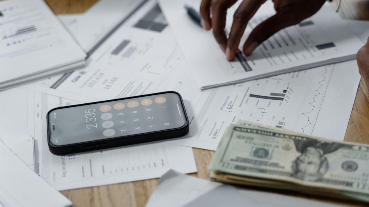 Close-up of hands calculating financial data with reports, smartphone, and cash on table.