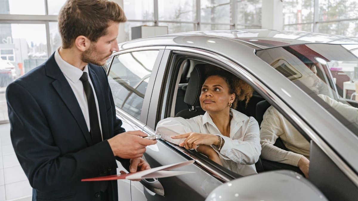 A car dealer hands keys to a woman sitting inside a vehicle in a car showroom.