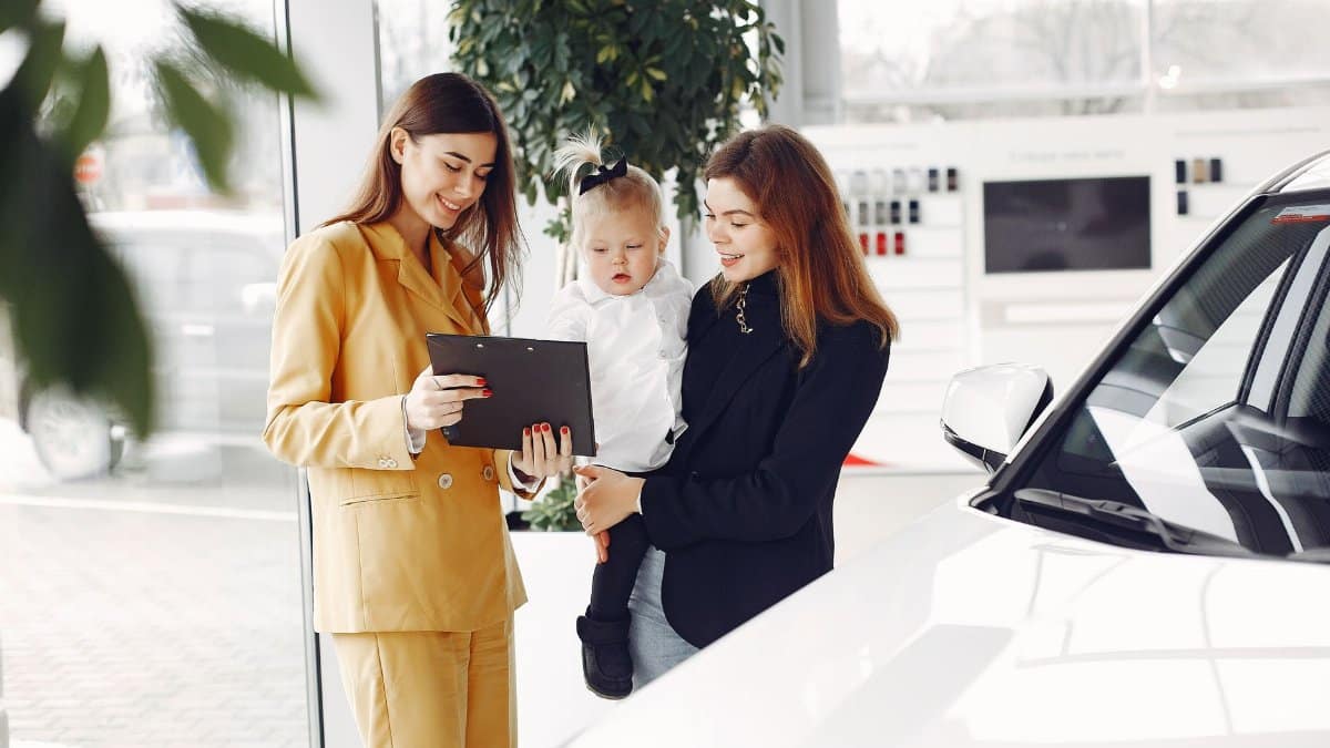 Positive young female with cute little daughter on hands discussing car characteristics with professional dealer in stylish beige suit while standing in car showroom in daylight
