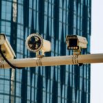 Surveillance cameras mounted on a pole in front of a modern glass building.