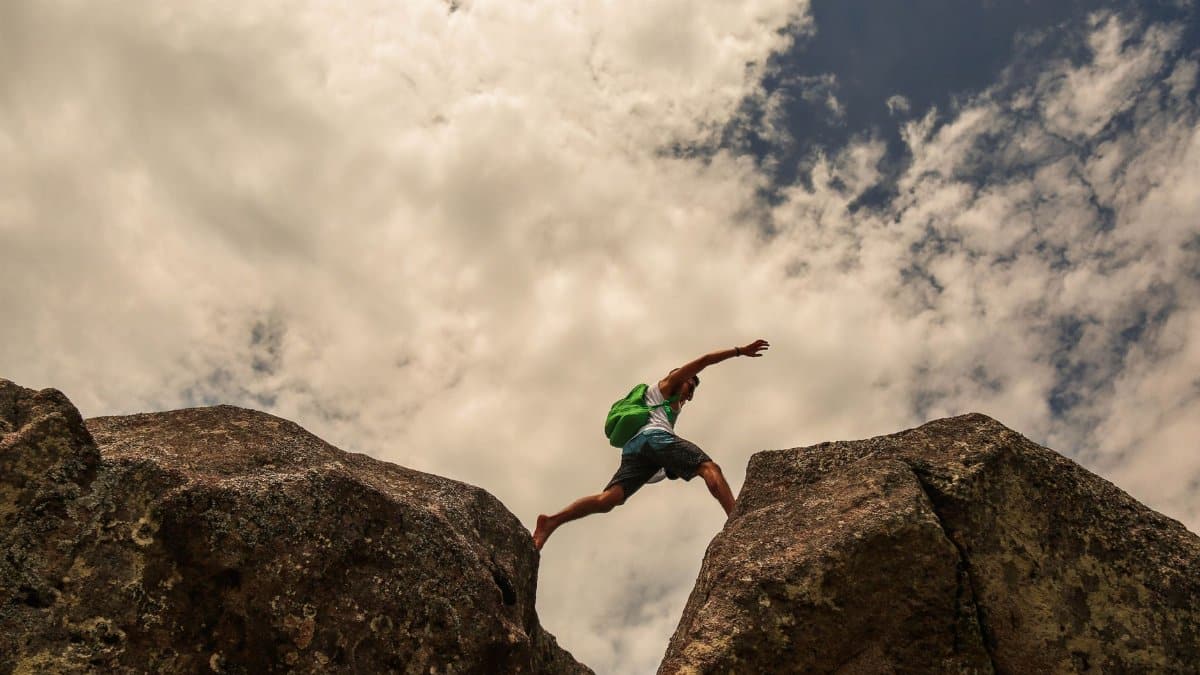 From below side view of fit anonymous traveler with backpack leaping bristly rocks under sky in clouds during trip