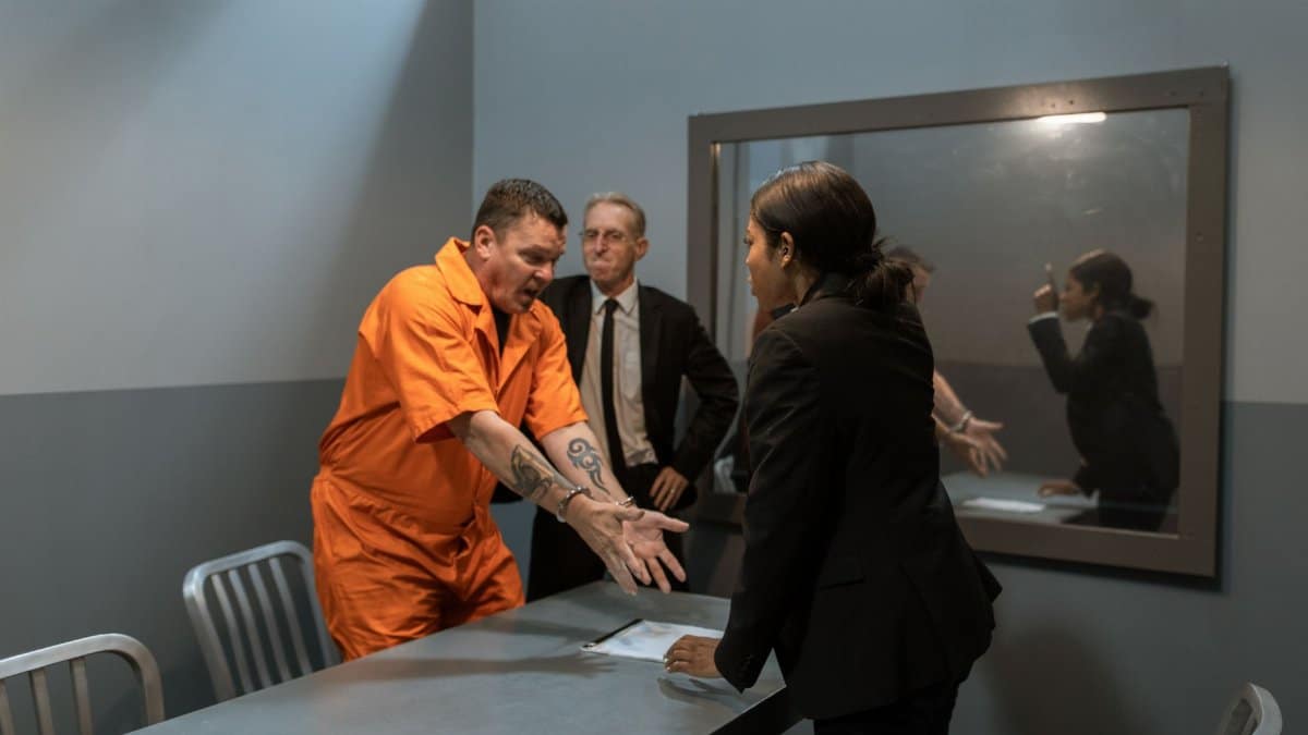 A prisoner in an orange jumpsuit discusses with officers in an interrogation room.