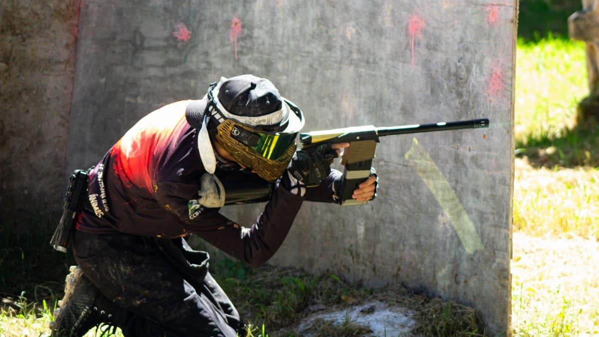 Energetic paintball player crouching with marker behind cover outdoors on a sunny day.