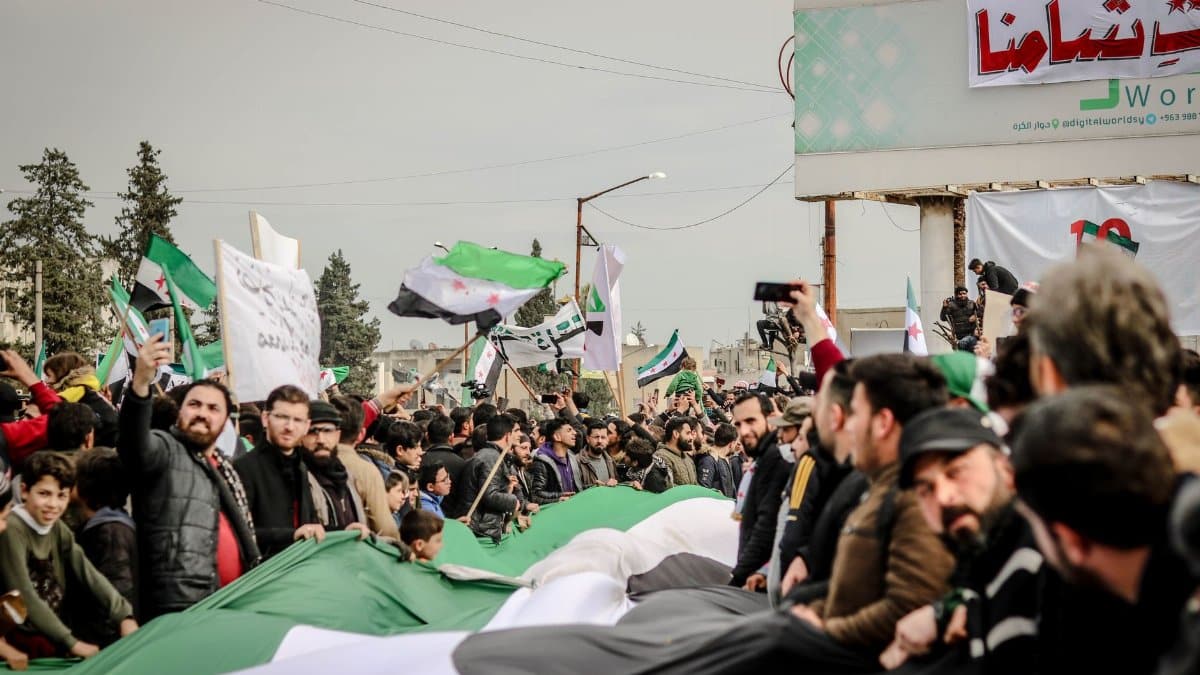 Crowds gather for a protest in Idlib, Syria, with flags and signs advocating political change.