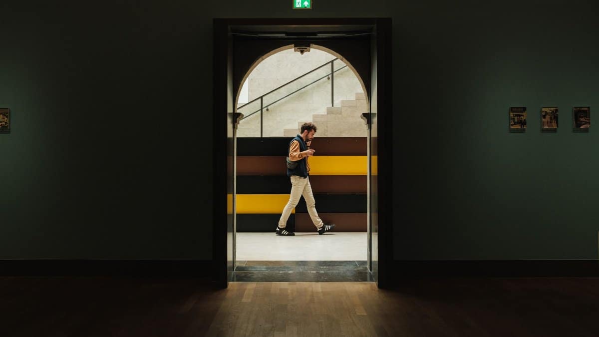 A man strolls through an art museum, framed by a doorway, with modern architecture in the background.