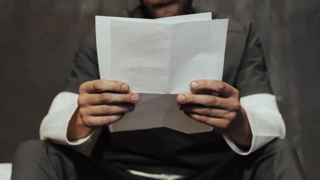 Close-up of a prisoner in uniform reading a letter in a cell setting, highlighting prison life.