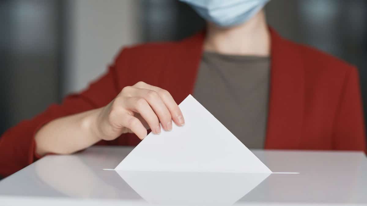 Person wearing a face mask dropping a ballot into a box to cast a vote. Emphasizes civic duty.