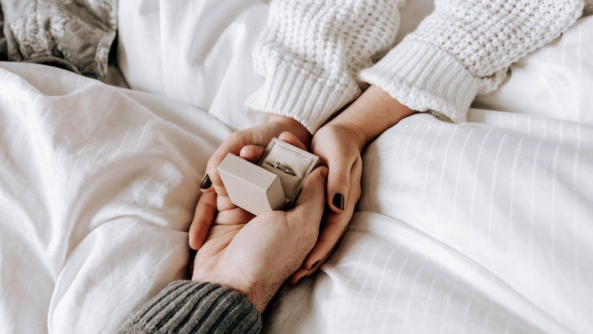 From above crop anonymous male making proposal and presenting engagement ring while lying together on cozy bed