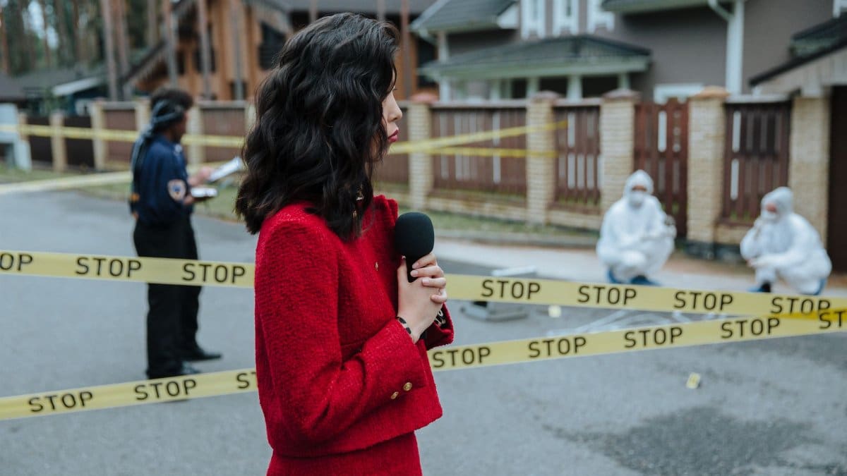 Female journalist reporting live on a crime scene investigation with police presence outdoors.