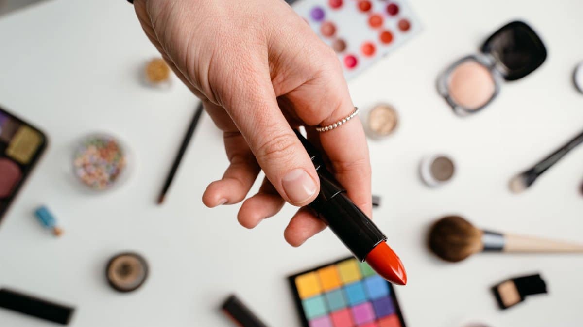 A hand holding a red lipstick surrounded by various makeup products on a light background.