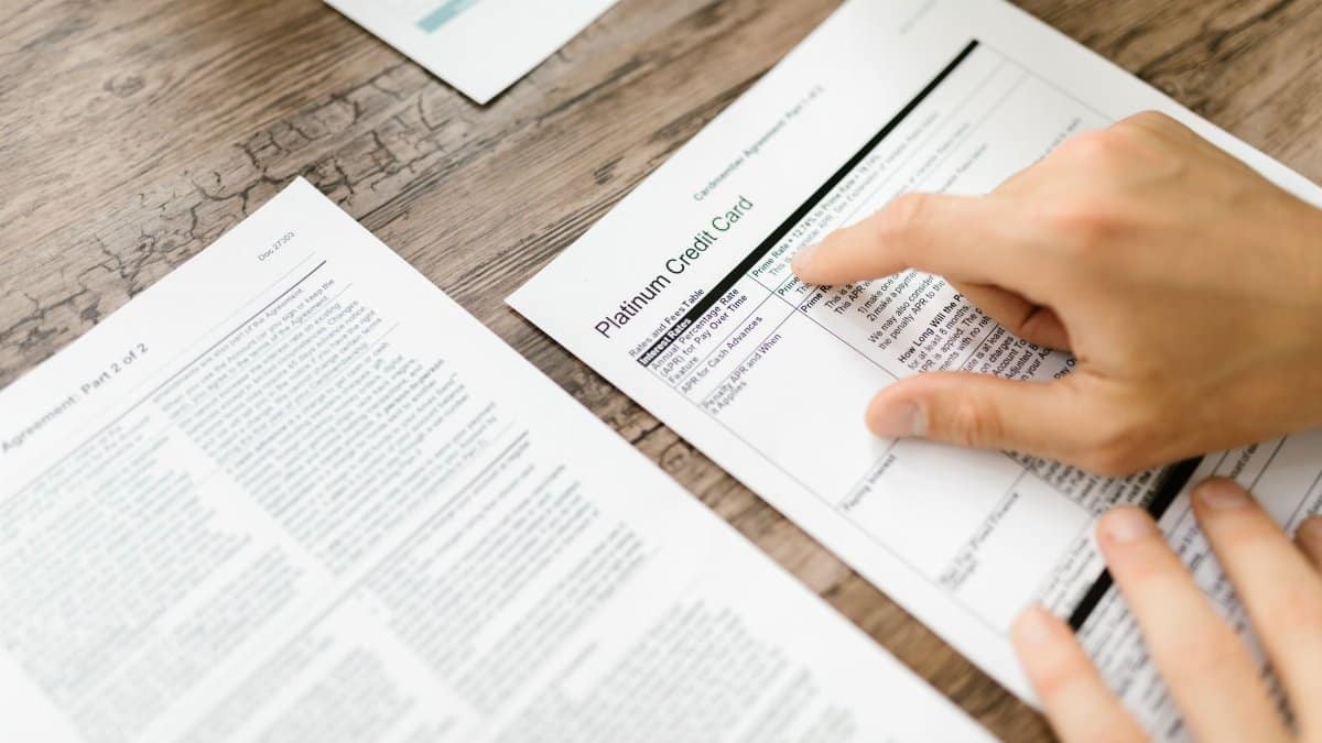 A hand examining a credit card agreement on a wooden desk, highlighting financial review.