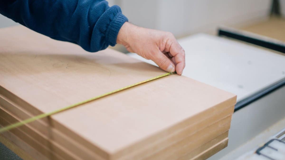 Close-up of a carpenter's hand using a tape measure to measure wooden planks indoors.