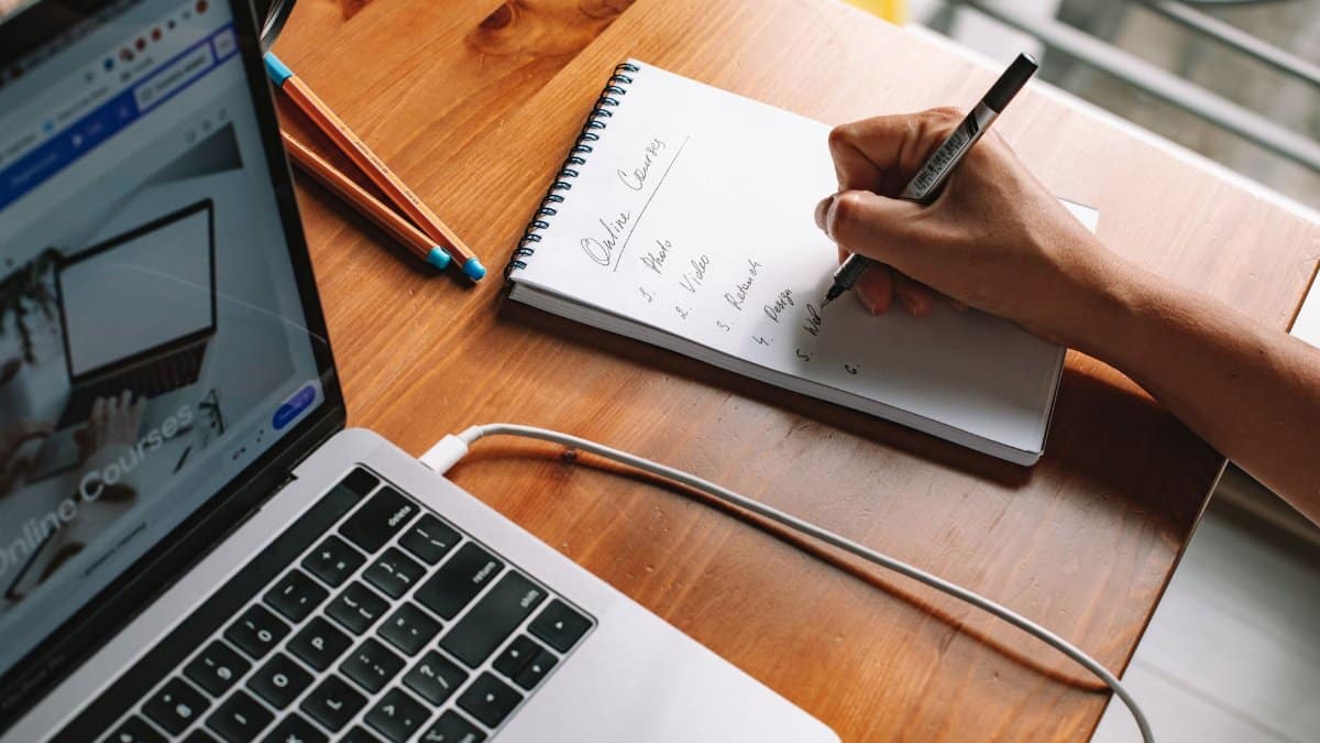 A person writing notes while researching online courses on a laptop in a home setting.