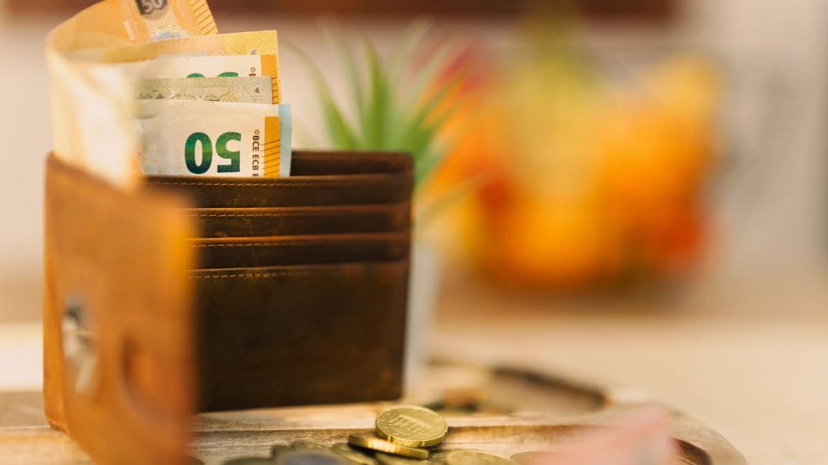 Brown wallet holding banknotes with scattered coins on a wooden table.
