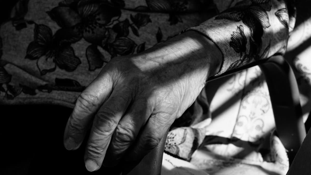 Black and white photo of an elderly hand resting on a chair, showing age and texture.