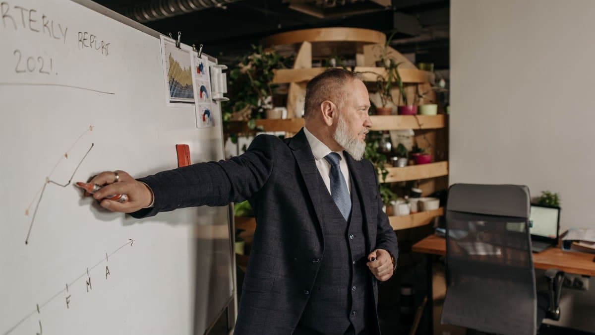 A businessman in a suit presents a quarterly report on a whiteboard in a modern office setting.